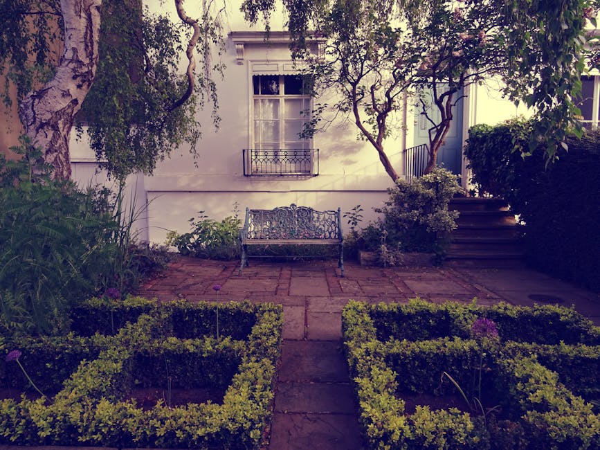 A small, well-maintained garden in front of a white house with a window and flower box, featuring neatly trimmed hedges forming geometric patterns, a paved stone pathway leading to a wrought iron benc