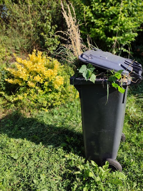 A black wheeled garden waste bin placed on a lush green lawn, filled with garden pruning debris including dried grass, leafy stems, and small branches. Surrounding the bin, there are various plants an