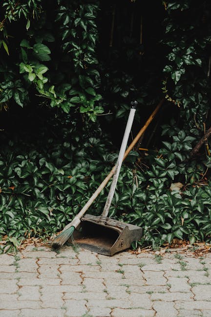 A black wheeled garden waste bin placed on a lush green lawn, filled with garden pruning debris including dried grass, leafy stems, and small branches. Surrounding the bin, there are various plants an
